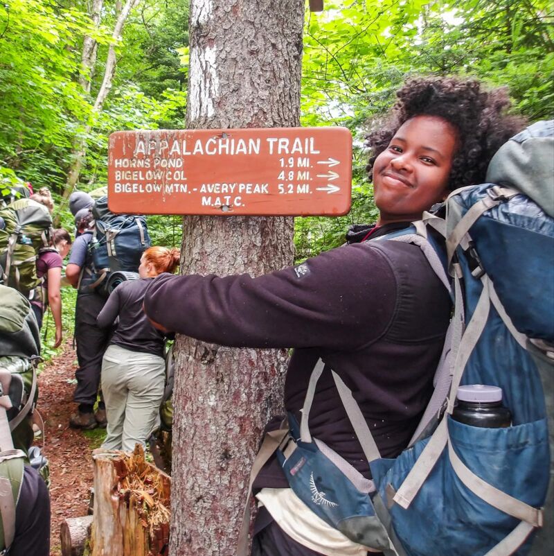 The image shows a woman standing next to a sign for the Appalachian Trail. The sign indicates that Horns Pond is 1.9 miles away, Bigelow Col is 4.9 miles away, and Bigelow Mtn - Avery Peak is 5.2 miles away. The woman is wearing a backpack and appears to be hiking. Other hikers are visible in the background, suggesting a group outing in a wooded area.